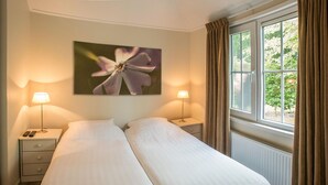 Un lit double avec du linge de lit blanc, deux tables de chevet avec des lampes, une fenêtre avec des rideaux et un tableau représentant une fleur accroché au mur.