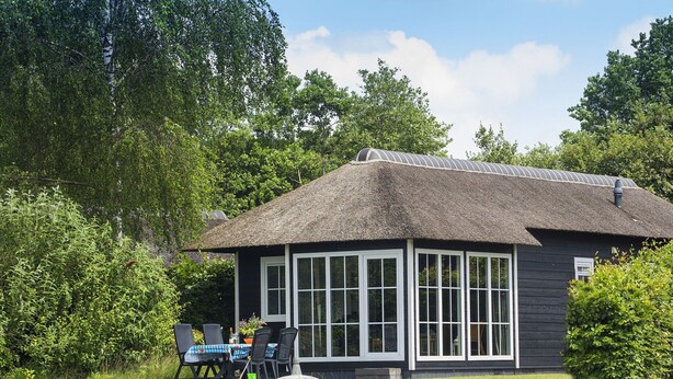 Un chalet au toit de chaume, doté d’un grand porche vitré, entouré d’une végétation luxuriante et bordé par un plan d’eau.