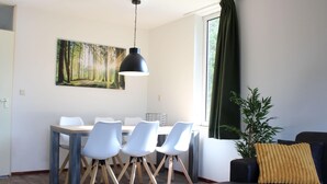 Un salon moderne avec un canapé noir, une table à manger avec des chaises blanches, un grand tableau représentant une forêt et un rideau vert.