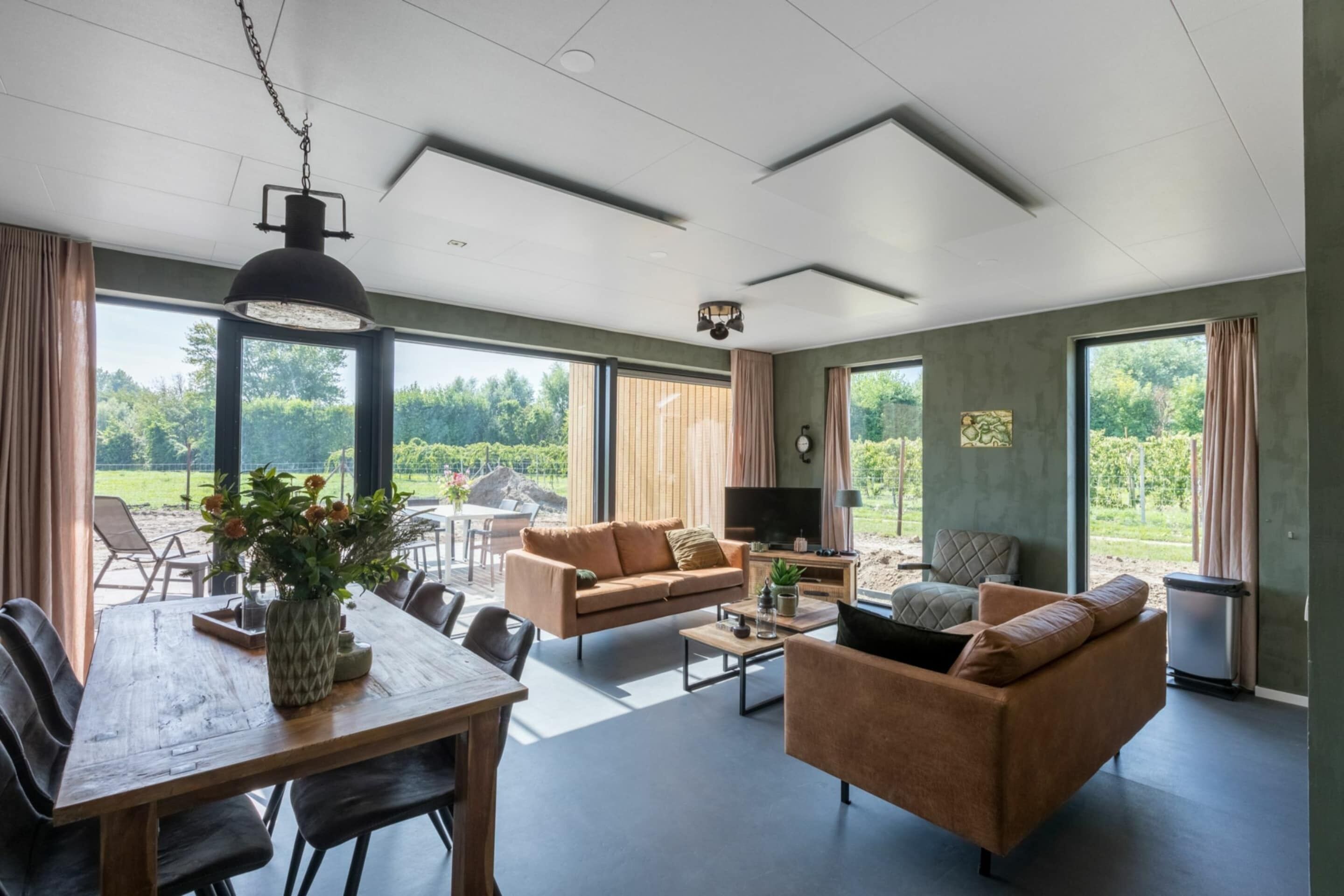Ein modernes Wohnzimmer mit Esstisch aus Holz, Sofas und einem Fernseher. Große Fenster bieten Blick ins Grüne.