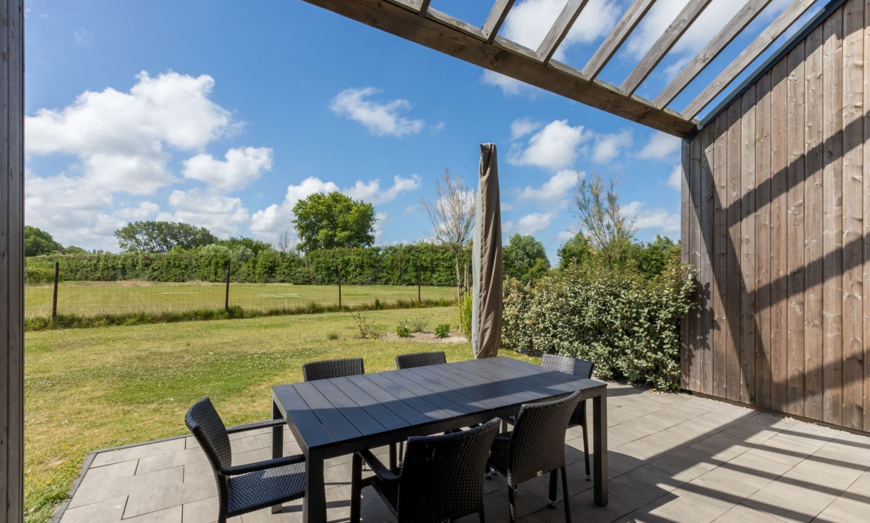 Un patio esterno con tavolo e sedie, una recinzione in legno e vista su un prato e alberi.