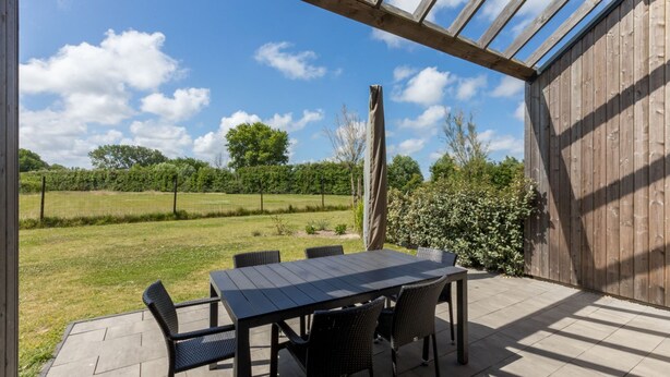 An outdoor patio with a table and chairs, a wooden fence, and a view of a grassy field and trees. - Holiday Home in Vrouwenpolder Near Beach (Vrouwenpolder)