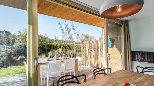 Un espace repas moderne avec une table et des chaises en bois, un saladier rempli de fruits et une vue sur un jardin et des arbres.