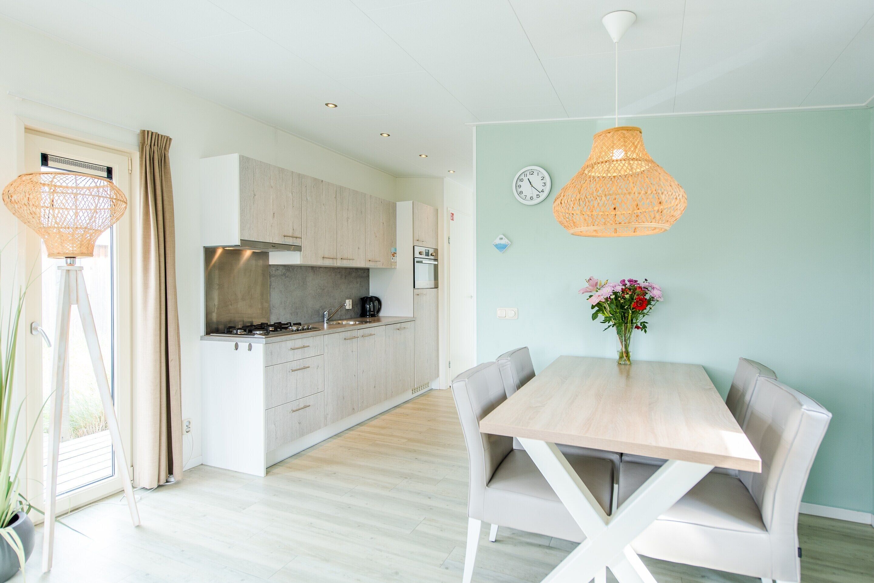 Une cuisine moderne avec des meubles de couleur bois clair, une hotte en acier inoxydable et un coin repas comprenant une table et des chaises en bois.