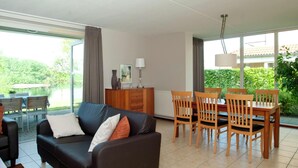 A living room with a black sofa, wooden dining table, and chairs. There is a window with curtains, a radiator, and a lamp on a cabinet. - Villa With Lake Views (Heel)