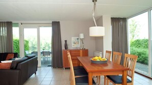 A modern living room with a sofa, dining table, and chairs. There is a window with curtains, a lamp, and a view of greenery outside. - Villa With Lake Views (Heel)