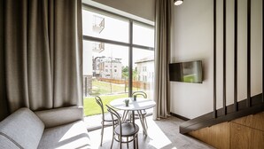 Una sala de estar moderna con un sofá, una mesa redonda de comedor con sillas, un televisor montado en la pared y grandes ventanales que ofrecen vistas al exterior.