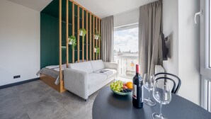 Una sala de estar moderna con un sofá, una mesa de comedor con un frutero y copas de vino, y vistas a un paisaje urbano a través de grandes ventanales.