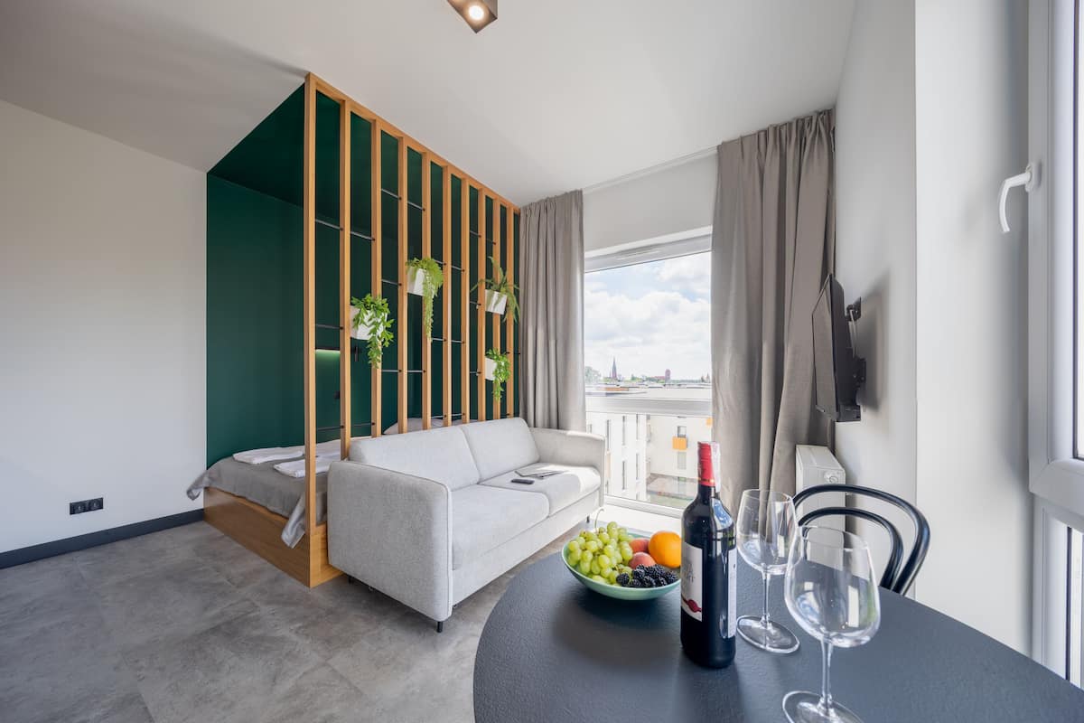Una sala de estar moderna con un sofá, una mesa de comedor con un frutero y copas de vino, y vistas a un paisaje urbano a través de grandes ventanales.