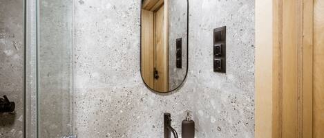 A bathroom with a glass shower enclosure, a white sink, a mirror, and a wooden door.