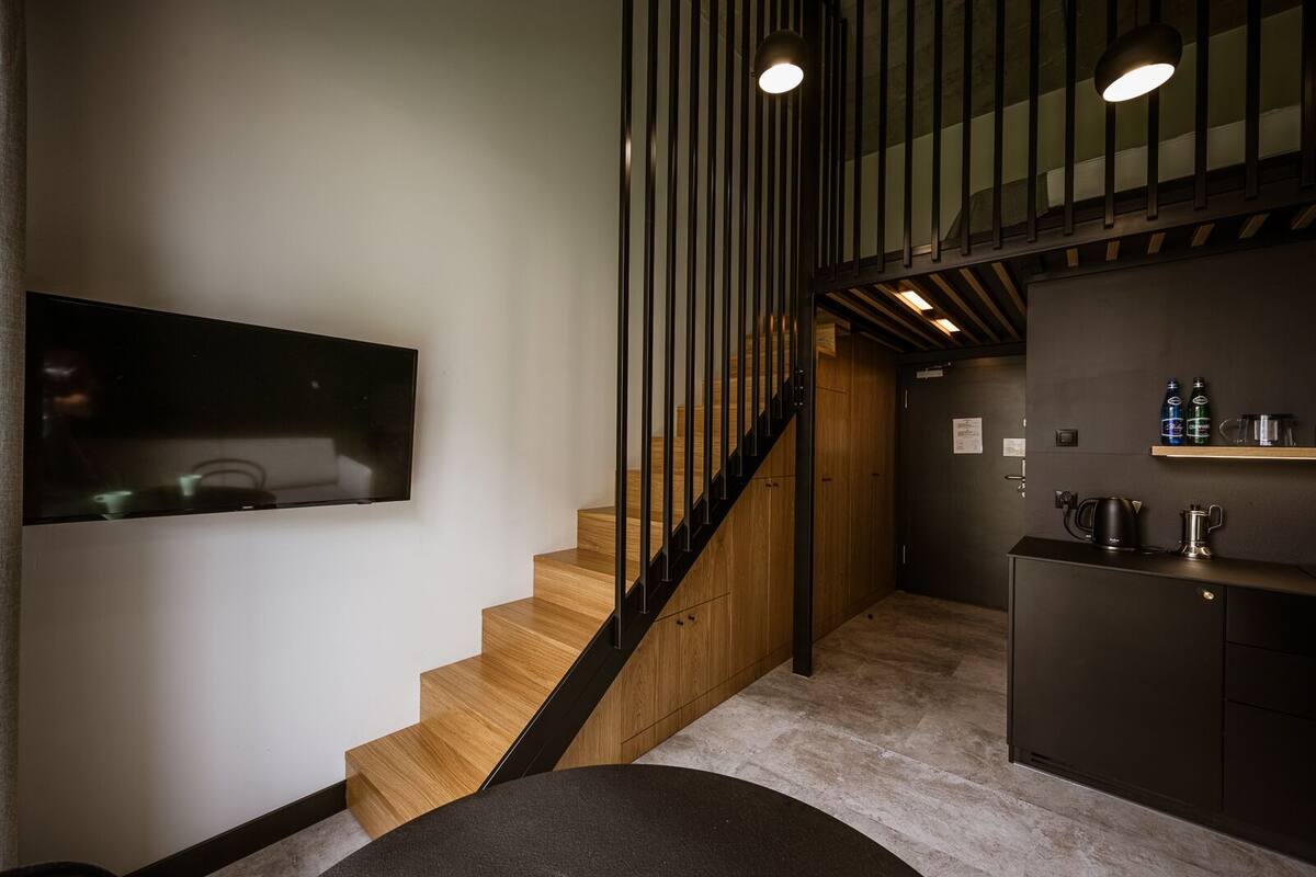 Una escalera moderna con peldaños de madera y barandillas negras, que conduce a una cocina de tonos oscuros con una cafetera y botellas en la encimera.