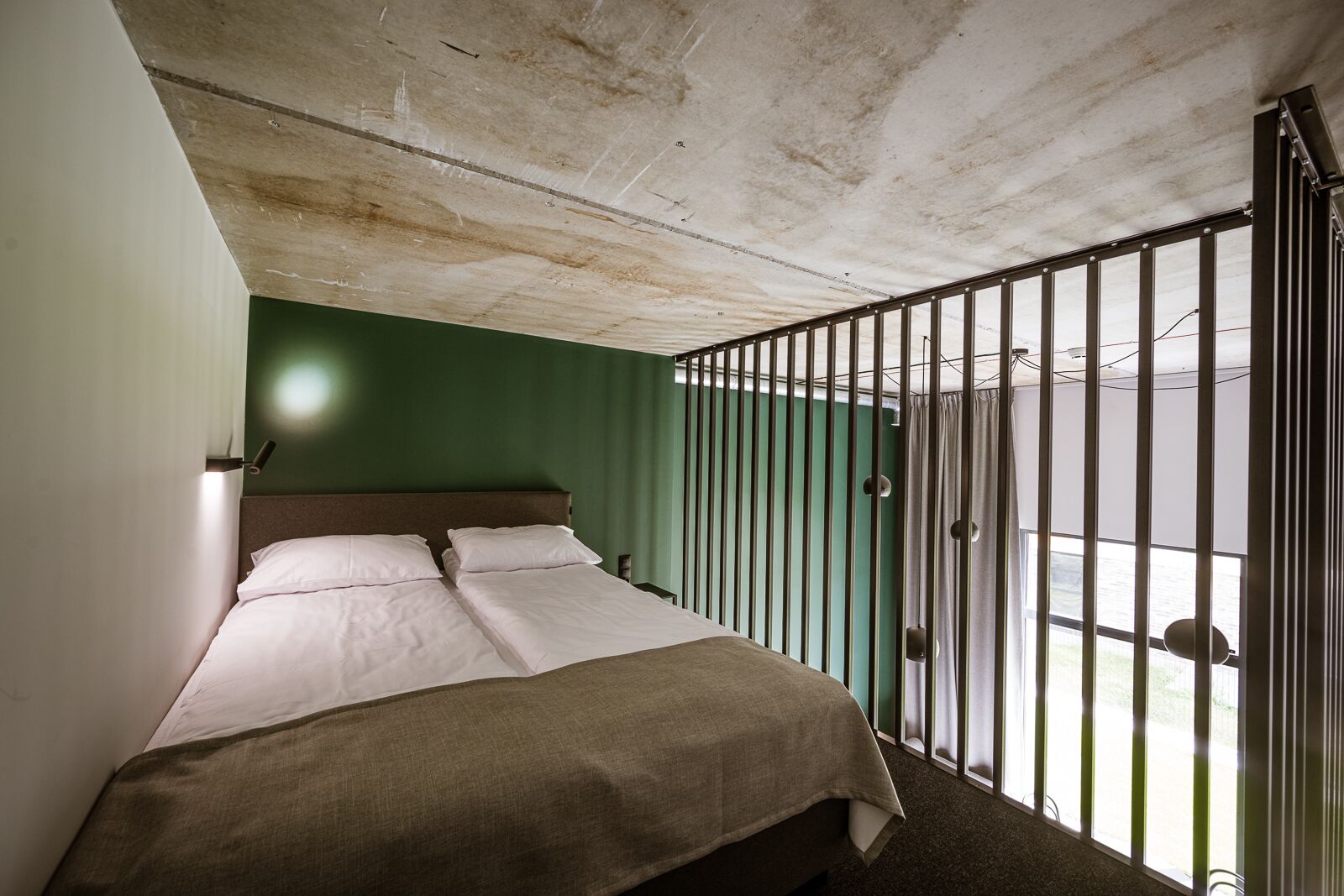 Uma cama de solteiro com cabeceira, uma janela com persianas verticais e teto de concreto.