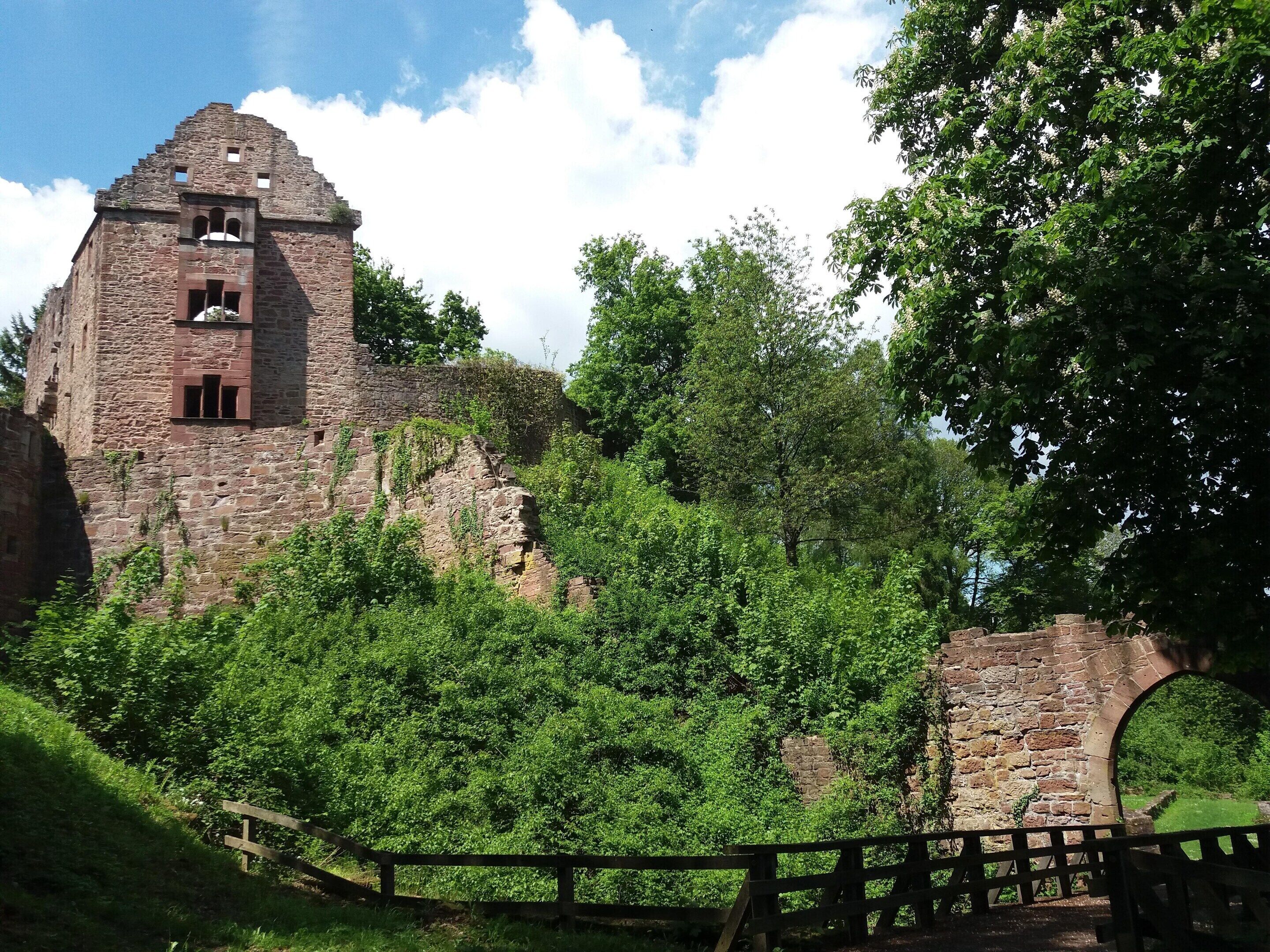 A historic stone building with a tower, surrounded by lush greenery and a wooden fence.