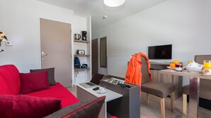 Ett modernt vardagsrum med en röd soffa, ett soffbord och en bärbar dator. Det finns en matplats med ett bord och stolar, och ett köksområde med en mikrovågsugn och en brödrost.