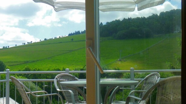 Ein Balkon mit Korbstühlen und einem Tisch, der auf ein grünes Feld mit Zaun und Bäumen blickt.