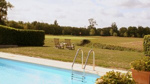 A swimming pool with a metal ladder, surrounded by a hedge, with chairs and a golf course in the background.