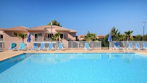 Piscina com espreguiçadeiras e guarda-sóis, cercada por uma vedação, com edifícios ao fundo.