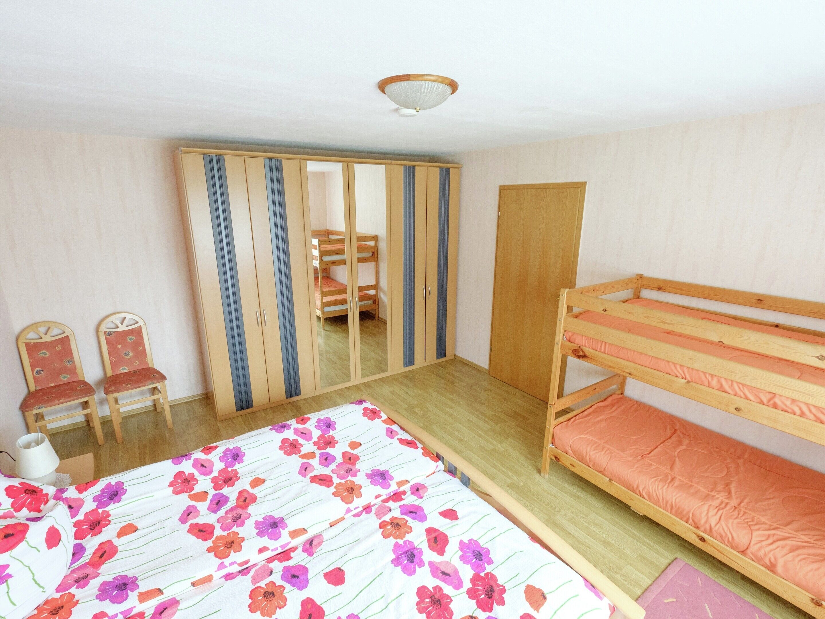 Une chambre avec un lit superposé, une armoire, une chaise et un lit recouvert d’une couvre-lit à motifs floraux.