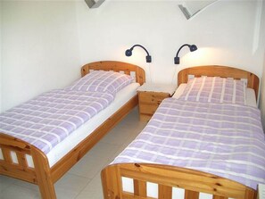 Deux lits simples avec des cadres et des têtes de lit en bois, chacun étant muni d’une table de chevet et d’une lampe fixée au mur.