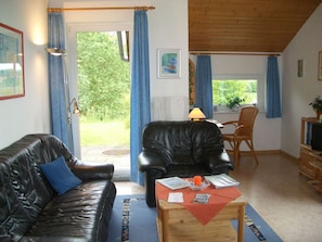 A living room with a leather sofa, a wooden coffee table, and a chair. There is a door leading to an outdoor area with a view of greenery. - Bungalow im Feriendorf Uslar mit Gartenterrasse (Uslar)