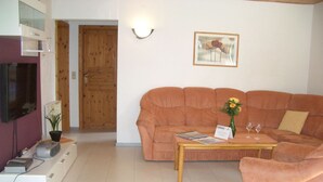 A living room with a sofa, a coffee table, a TV, and a door. - Bungalow im Feriendorf Uslar mit Gartenterrasse (Uslar)