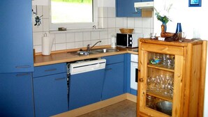A kitchen with blue cabinets, a microwave, a sink, and a wooden cabinet with glassware. - Bungalow im Feriendorf Uslar mit Gartenterrasse (Uslar)