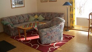A living room with a sofa, armchair, coffee table, and a lamp. There are two framed pictures on the wall and a window with curtains. - Bungalow im Feriendorf Uslar mit Gartenterrasse (Uslar)