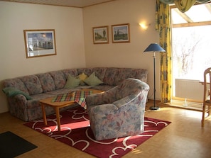 Una sala de estar con sofá, sillón, mesa de centro y una lámpara. Hay dos cuadros en la pared y una ventana con cortinas.