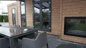 Un área de comedor exterior moderna con pared de madera y puertas de vidrio, una mesa con sillas y un hogar para fuego.