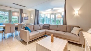A modern living room with a beige sectional sofa, a coffee table, and a dining area with chairs and a table. - Restyled Thatched Villa Near the Sea in Cosy Domburg (Domburg)