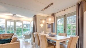 A spacious dining area with a long table and multiple chairs, large windows offering a view of greenery, and a modern ceiling light fixture. - Restyled Thatched Villa Near the Sea in Cosy Domburg (Domburg)