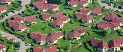 An aerial view of a residential area with multiple houses, each with a red roof, surrounded by well-maintained lawns and parked cars.