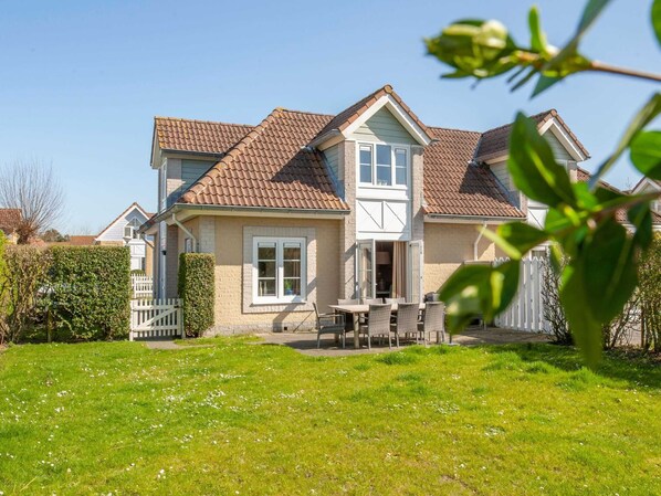 A two-story house with a tiled roof, a front porch with a table and chairs, and a well-maintained lawn. - Semi-detached Villa With Microwave Near the Beach (Kamperland)