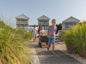 Stroll along the paved pathway to our beachfront property, surrounded by lush grasses and leading to modern buildings with balconies.