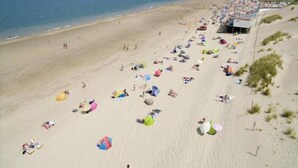 Una spiaggia con persone che prendono il sole e nuotano, tende e un molo sullo sfondo.