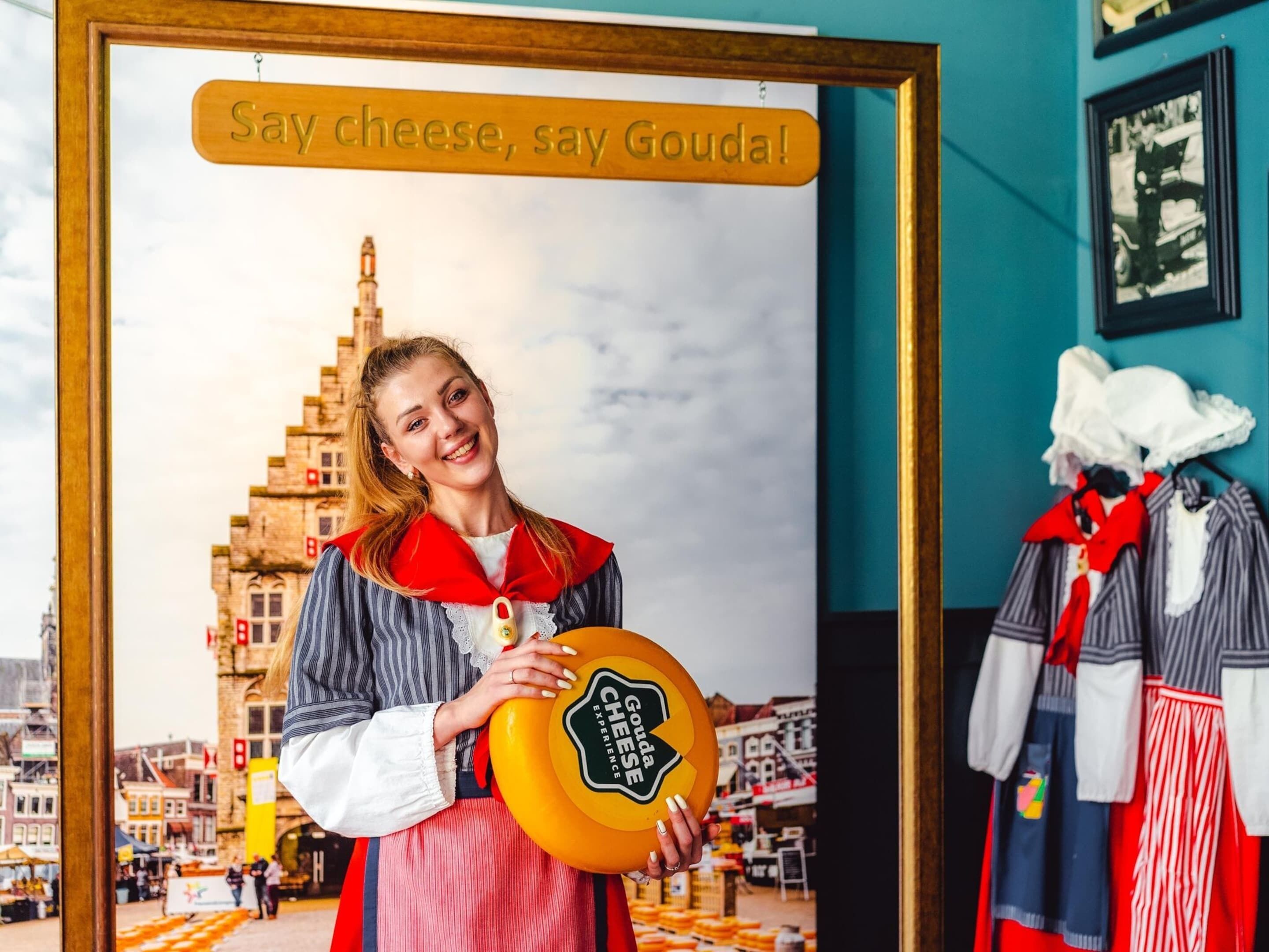 Una persona che tiene in mano un cartello giallo con su scritto 'Say cheese, say Gouda!', in piedi di fronte a una cornice con uno sfondo urbano.