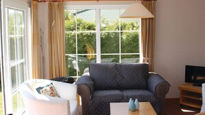 A living room with a sofa, a coffee table, a TV stand, and large windows with curtains. - Nicely Furnished Chalet with Oven near Wadden Sea (Tzummarum)