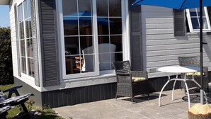 A house with a covered patio area featuring a table and chairs, a blue umbrella, and a window with shutters. - Nicely Furnished Chalet with Oven near Wadden Sea (Tzummarum)