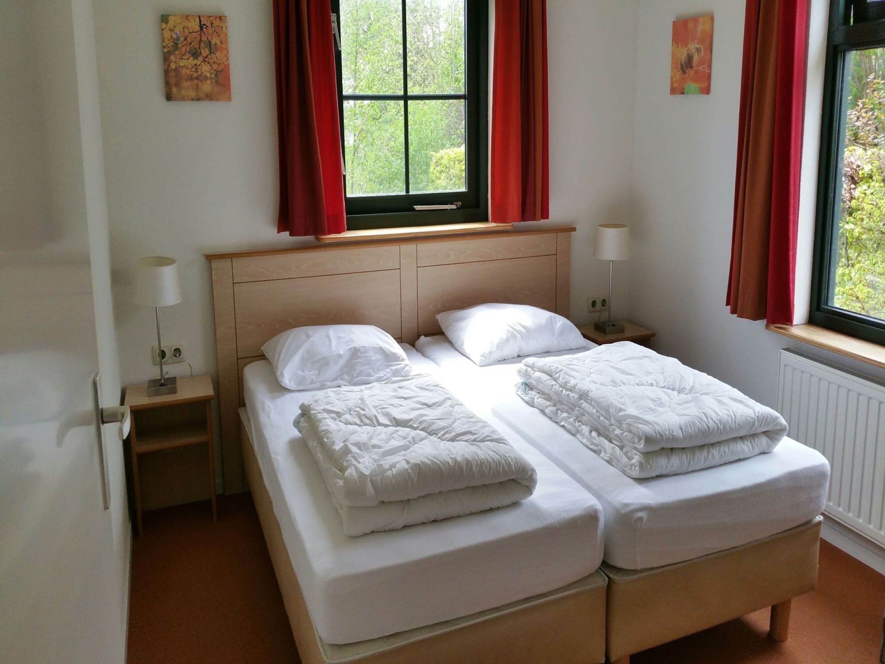 Una cama doble con ropa de cama blanca, cabecera de madera, dos mesitas de noche con lámparas, una ventana con cortinas rojas y un radiador.