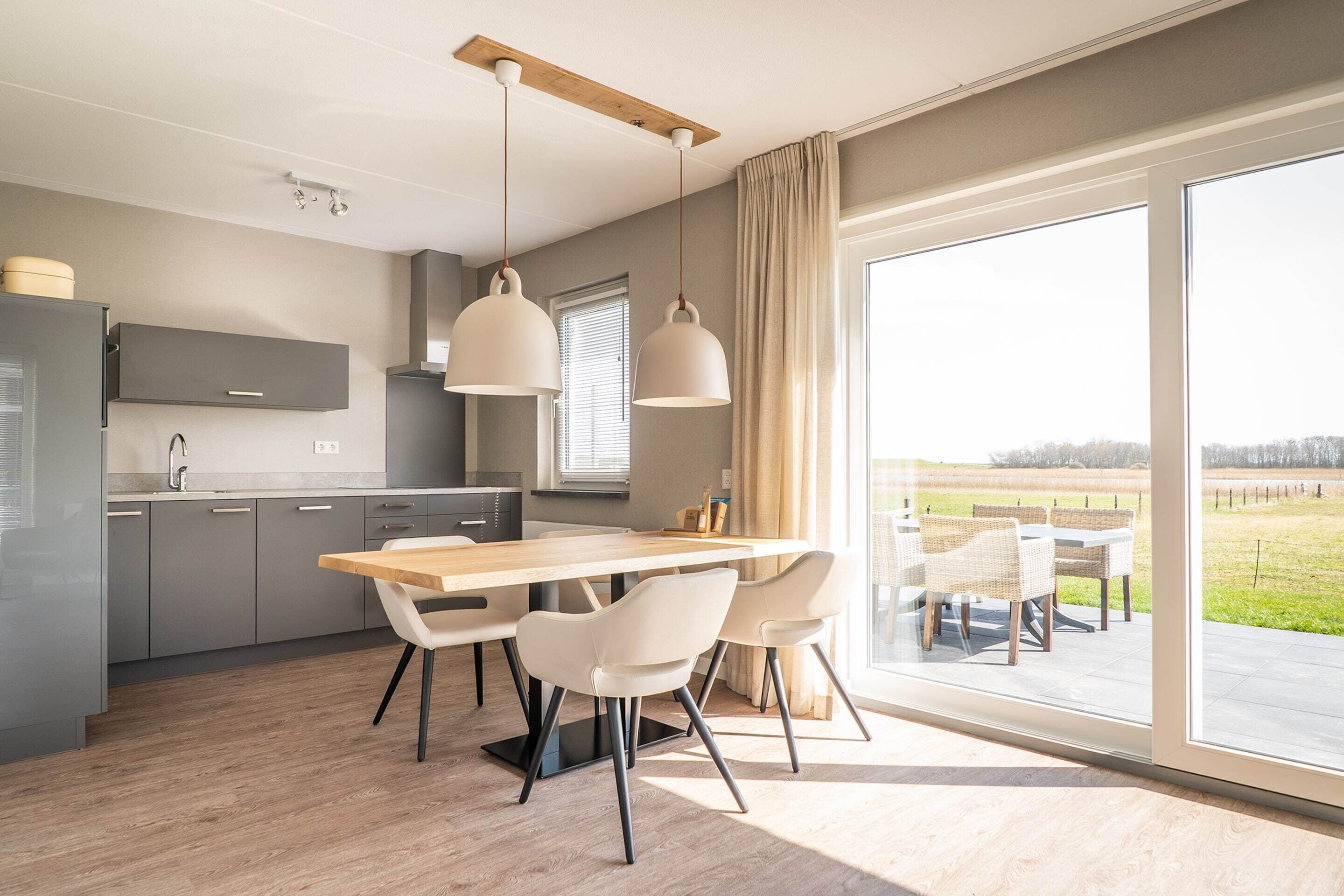A modern kitchen with grey cabinets, a wooden table, and white chairs. There is a large window with a view of a green landscape.