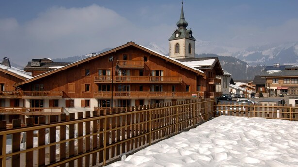 Un edificio de madera con balcones, una iglesia con aguja y suelo cubierto de nieve.