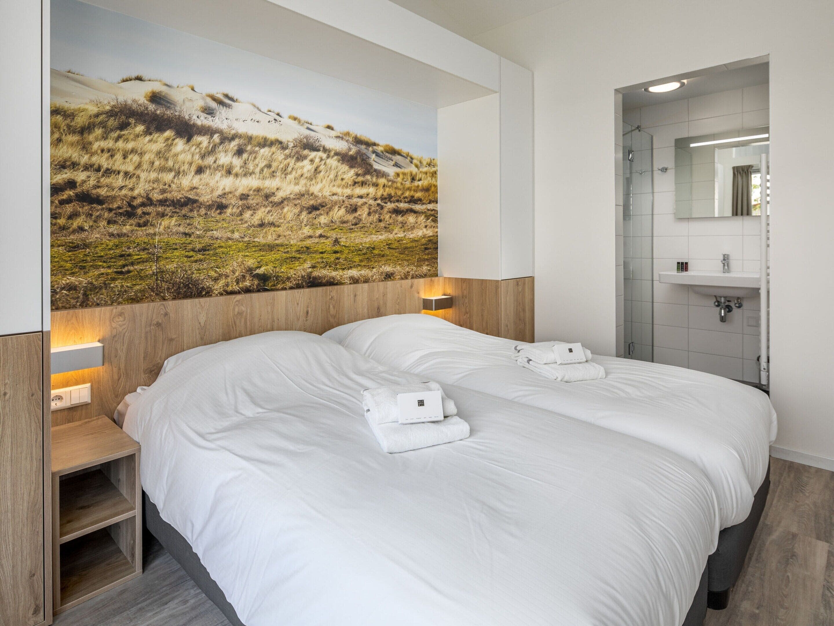 A modern hotel room with a large bed, two bedside tables, a wall-mounted TV, and a bathroom with a shower and sink.