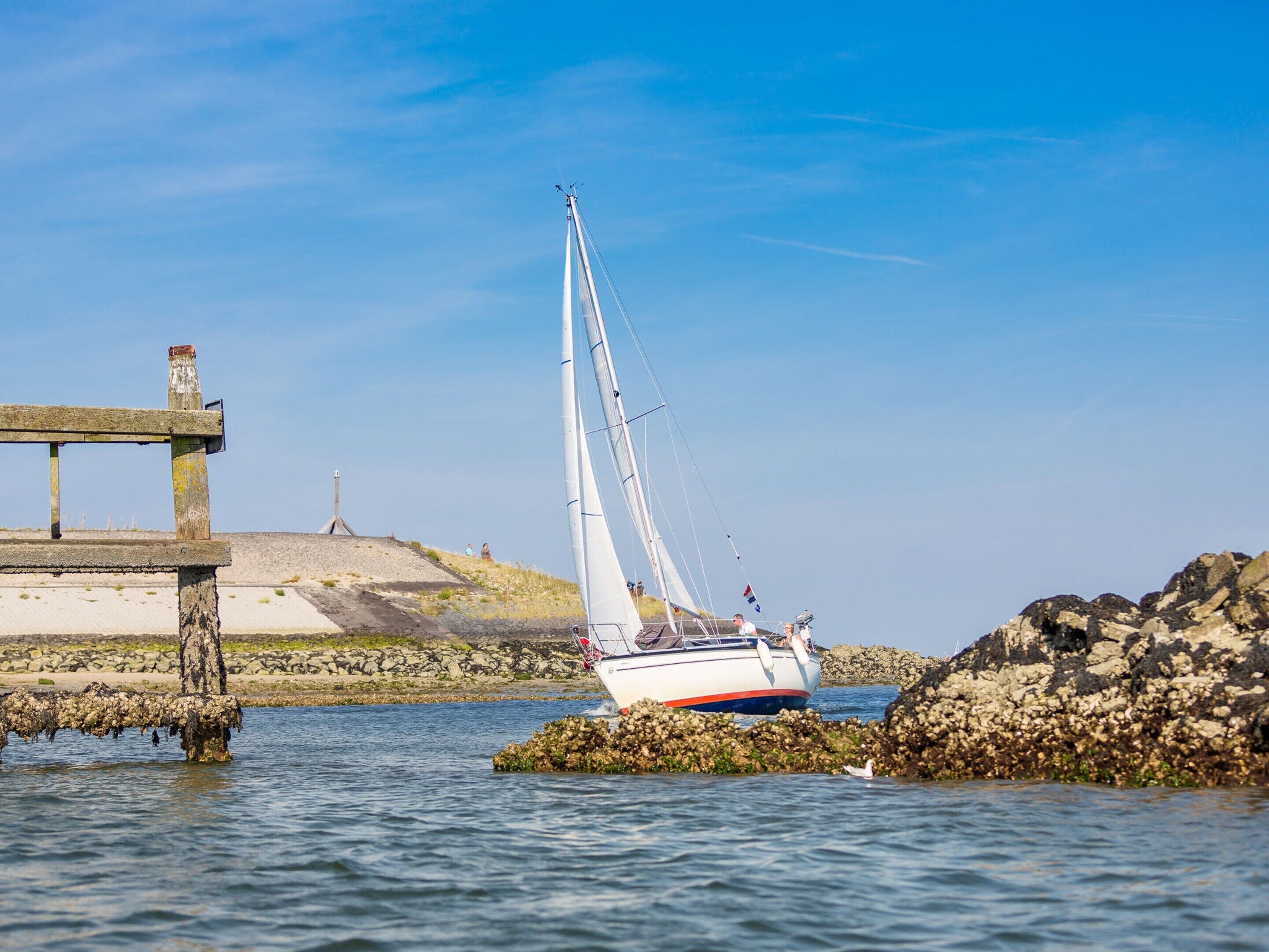 A sailboat anchored near a rocky shore with a wooden structure in the background.