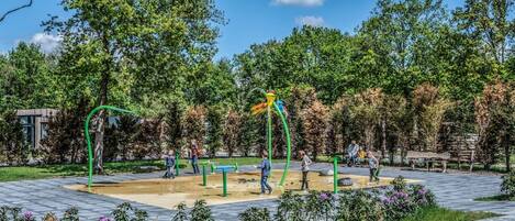 An outdoor playground with green exercise equipment, surrounded by trees and a paved area.