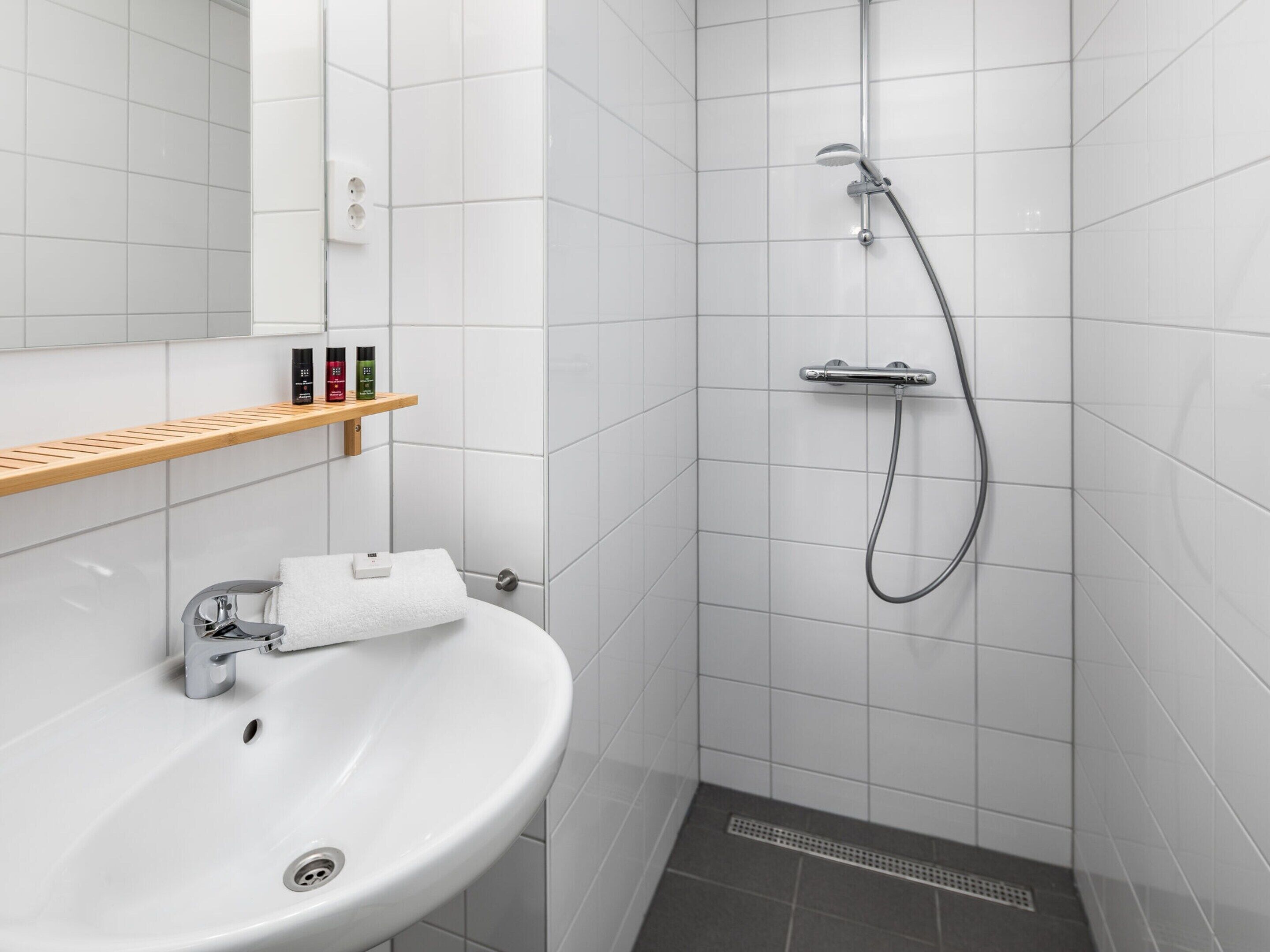 A bathroom with a white sink, a mirror, a shower with a handheld showerhead, and a shelf with toiletries.