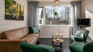 Ein modernes Wohnzimmer mit einem braunen Sofa, grünen Sesseln, einem kleinen runden Tisch mit einer Flasche und Gläsern sowie einem großen Fenster mit Blick auf Bäume.