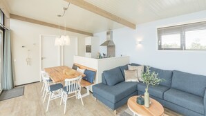 A modern living room with a sofa, dining table, and chairs, a kitchen area, and a large window with blinds. - Great Lodge with Microwave Oven near Beach (Hollum)