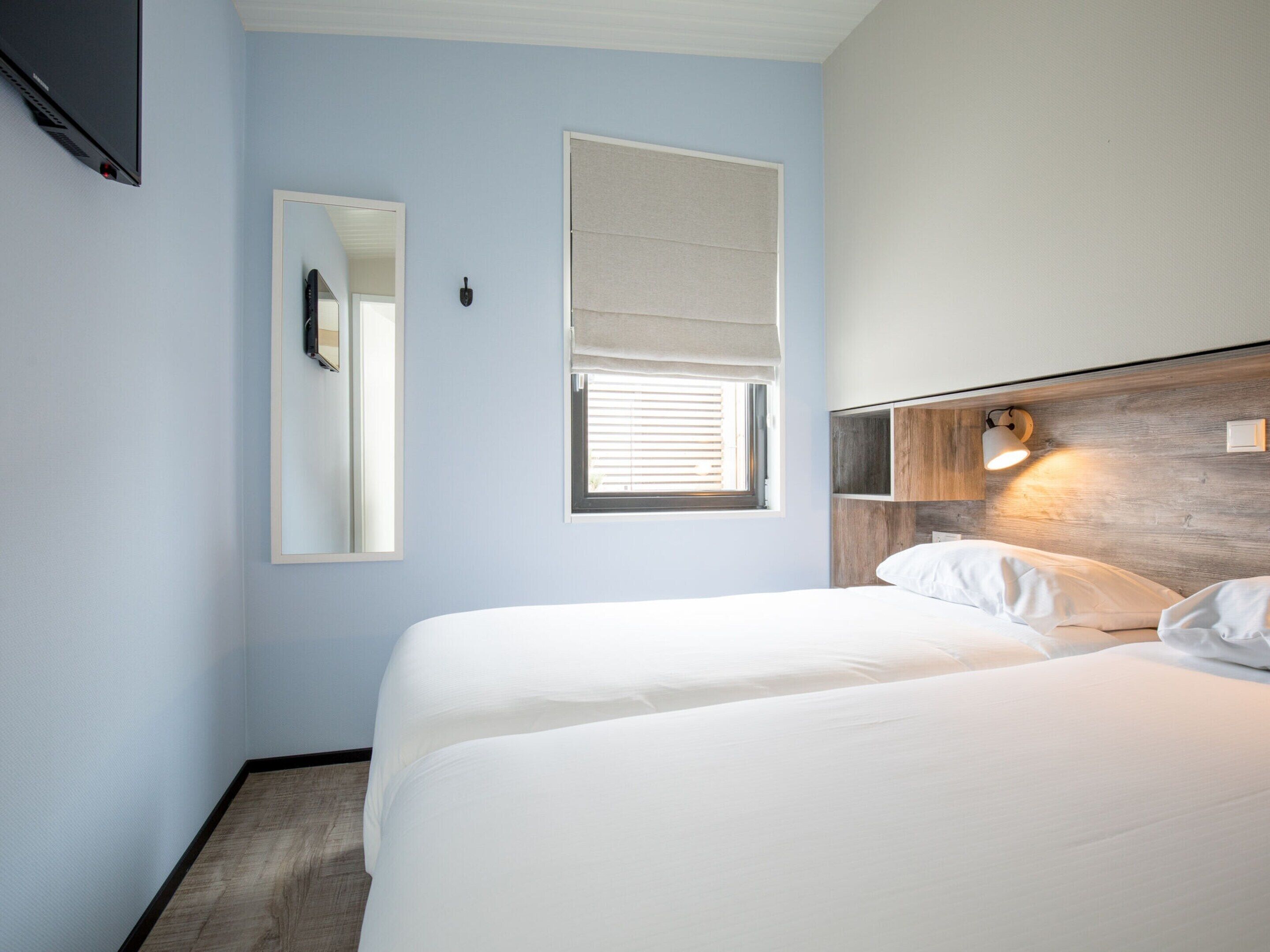 A hotel room with a bed, a mirror, a window with blinds, and a wall-mounted light fixture.
