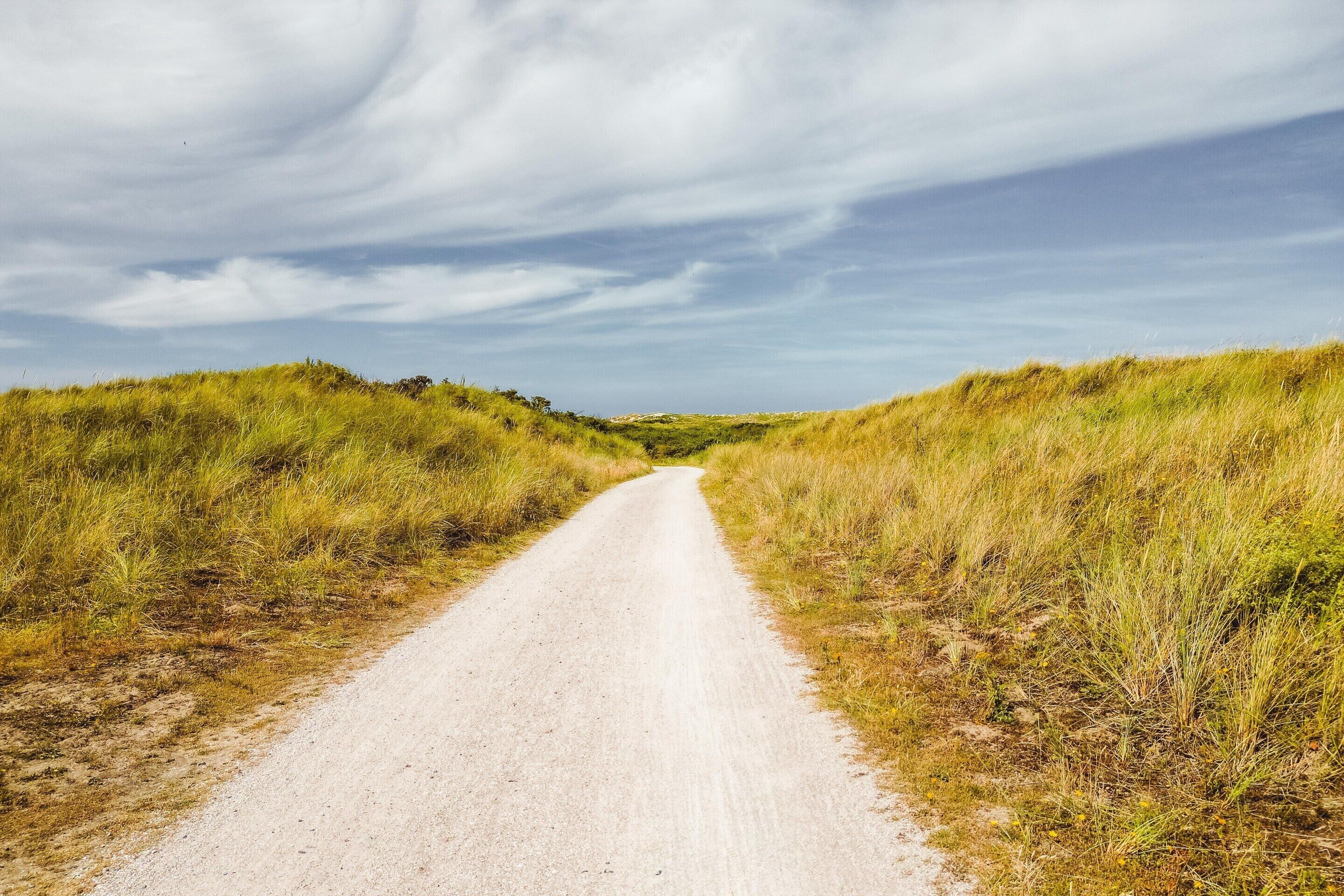 A rural dirt road flanked by grassy fields under a cloudy sky.