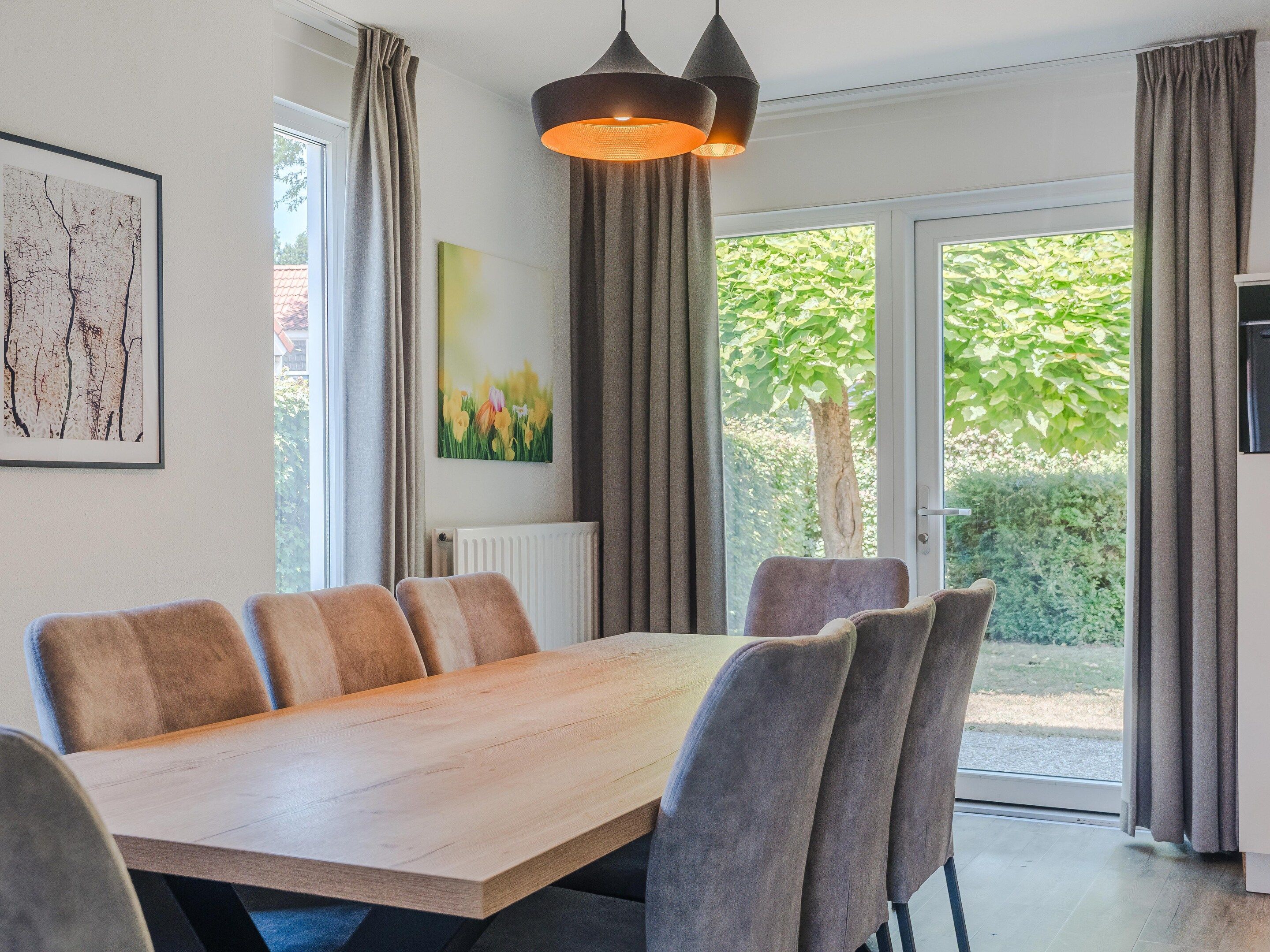 Uma área de jantar com mesa de madeira e cadeiras acolchoadas, uma luminária pendente e vista para o verde lá fora.